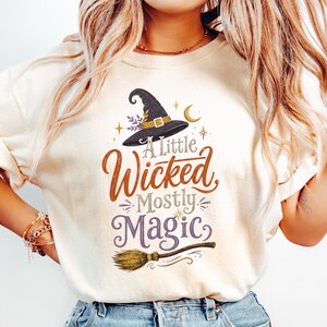 May include: A cream-colored t-shirt featuring a graphic of a black witch hat, a crescent moon, and a broomstick. The text reads A Little Wicked, Mostly MAGIC in orange, grey, and purple script and serif fonts with small star accents.