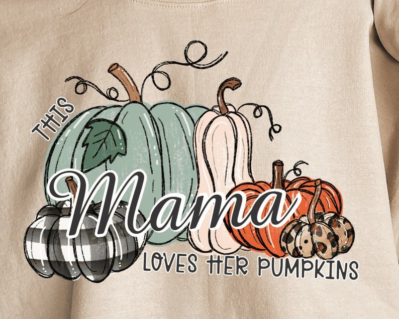 Pode incluir: Camisola bege com o gr&aacute;fico "This Mama loves her pumpkins". O gr&aacute;fico apresenta cinco ab&oacute;boras em v&aacute;rias cores e padr&otilde;es, incluindo verde, branco, laranja, preto e marrom.