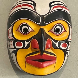 K&ouml;nnte beinhalten: Eine Holzmaske mit einem stilisierten menschlichen Gesicht, bemalt in Gelb, Rot, Schwarz und Wei&szlig;. Die Maske hat eine gro&szlig;e, prominente Nase und gro&szlig;e Augen.