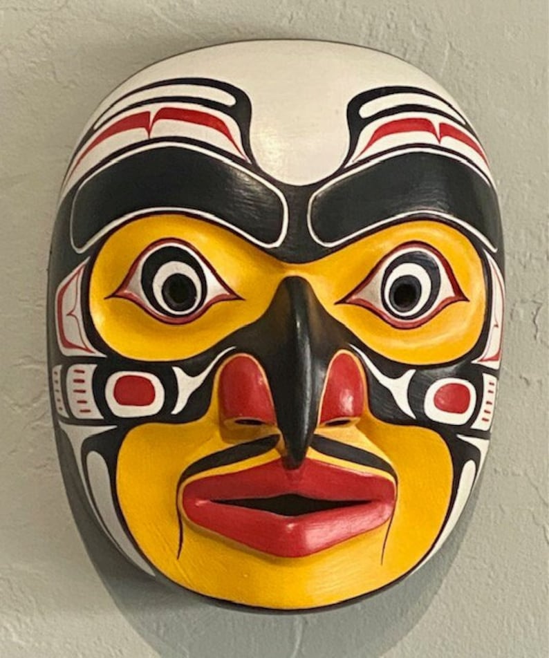 K&ouml;nnte beinhalten: Eine Holzmaske mit einem stilisierten menschlichen Gesicht, bemalt in Gelb, Rot, Schwarz und Wei&szlig;. Die Maske hat eine gro&szlig;e, prominente Nase und gro&szlig;e Augen.