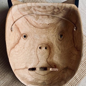 May include: A wooden mask with a carved face, featuring two eyes, a nose, and a mouth. The mask is painted in a natural wood color and has a black rim around the edges.