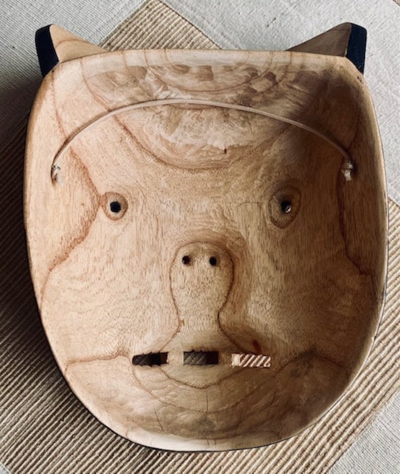 May include: A wooden mask with a carved face, featuring two eyes, a nose, and a mouth. The mask is painted in a natural wood color and has a black rim around the edges.