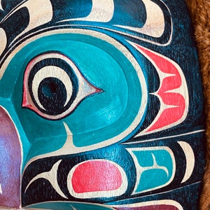 May include: Close-up of a carved wooden mask with a teal blue eye and red and white details. The mask is painted in a traditional style with black, white, teal blue, and red colors.