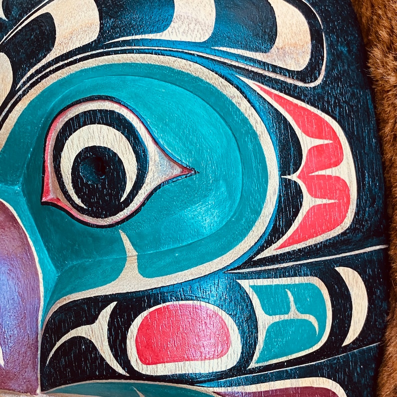 May include: Close-up of a carved wooden mask with a teal blue eye and red and white details. The mask is painted in a traditional style with black, white, teal blue, and red colors.
