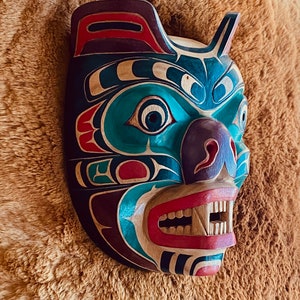 May include: A wooden bear mask with intricate carvings and vibrant colors, including teal, red, purple, and black. The mask has a fierce expression with sharp teeth and a wide open mouth.