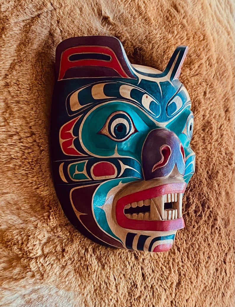 May include: A wooden bear mask with intricate carvings and vibrant colors, including teal, red, purple, and black. The mask has a fierce expression with sharp teeth and a wide open mouth.