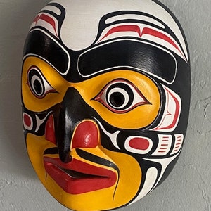 K&ouml;nnte beinhalten: Eine Holzmaske mit einem stilisierten menschlichen Gesicht, das in Schwarz, Wei&szlig;, Rot und Gelb bemalt ist. Die Maske hat eine gro&szlig;e, hervorstehende Nase und Augen sowie einen breiten Mund.