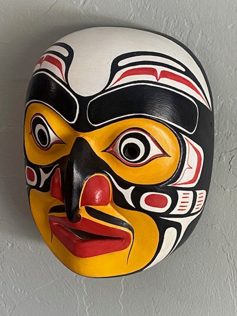 K&ouml;nnte beinhalten: Eine Holzmaske mit einem stilisierten menschlichen Gesicht, das in Schwarz, Wei&szlig;, Rot und Gelb bemalt ist. Die Maske hat eine gro&szlig;e, hervorstehende Nase und Augen sowie einen breiten Mund.