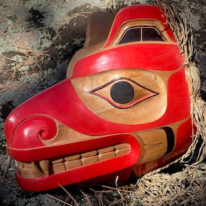 Peut inclure: Un masque en bois sculpté à la main représentant une tête d'animal stylisée, principalement rouge avec des accents en bois naturel. Le masque présente un design détaillé avec un œil proéminent, des dents et une surface texturée. Il s'agit probablement d'un artefact culturel ou d'une pièce décorative.