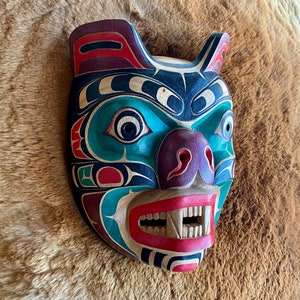 May include: A wooden carved mask depicting a bear with a green, blue, red, black, and purple paint design. The mask has a wide mouth with sharp teeth and a large nose.