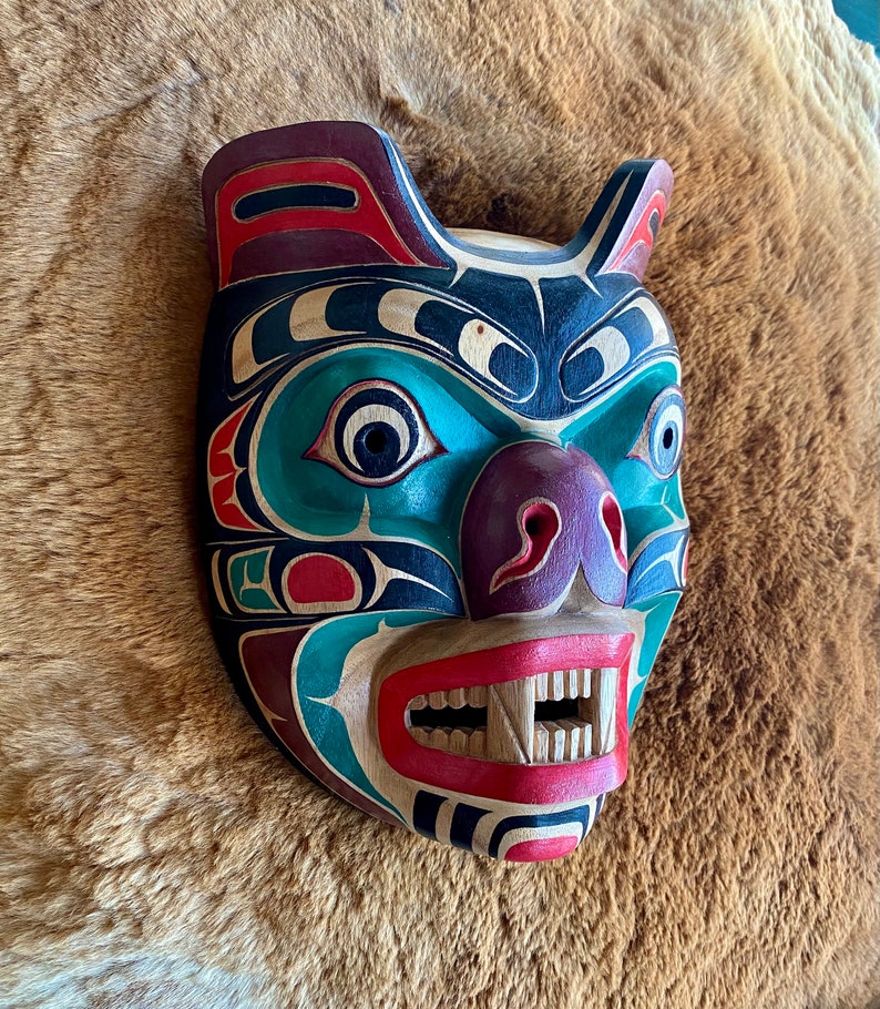May include: A wooden carved mask depicting a bear with a green, blue, red, black, and purple paint design. The mask has a wide mouth with sharp teeth and a large nose.