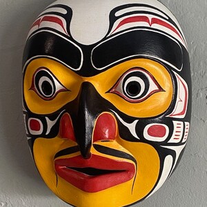 K&ouml;nnte beinhalten: Eine Holzmaske mit einem stilisierten menschlichen Gesicht, das in Schwarz, Wei&szlig;, Rot und Gelb bemalt ist. Die Maske hat eine gro&szlig;e, prominente Nase und gro&szlig;e Augen.
