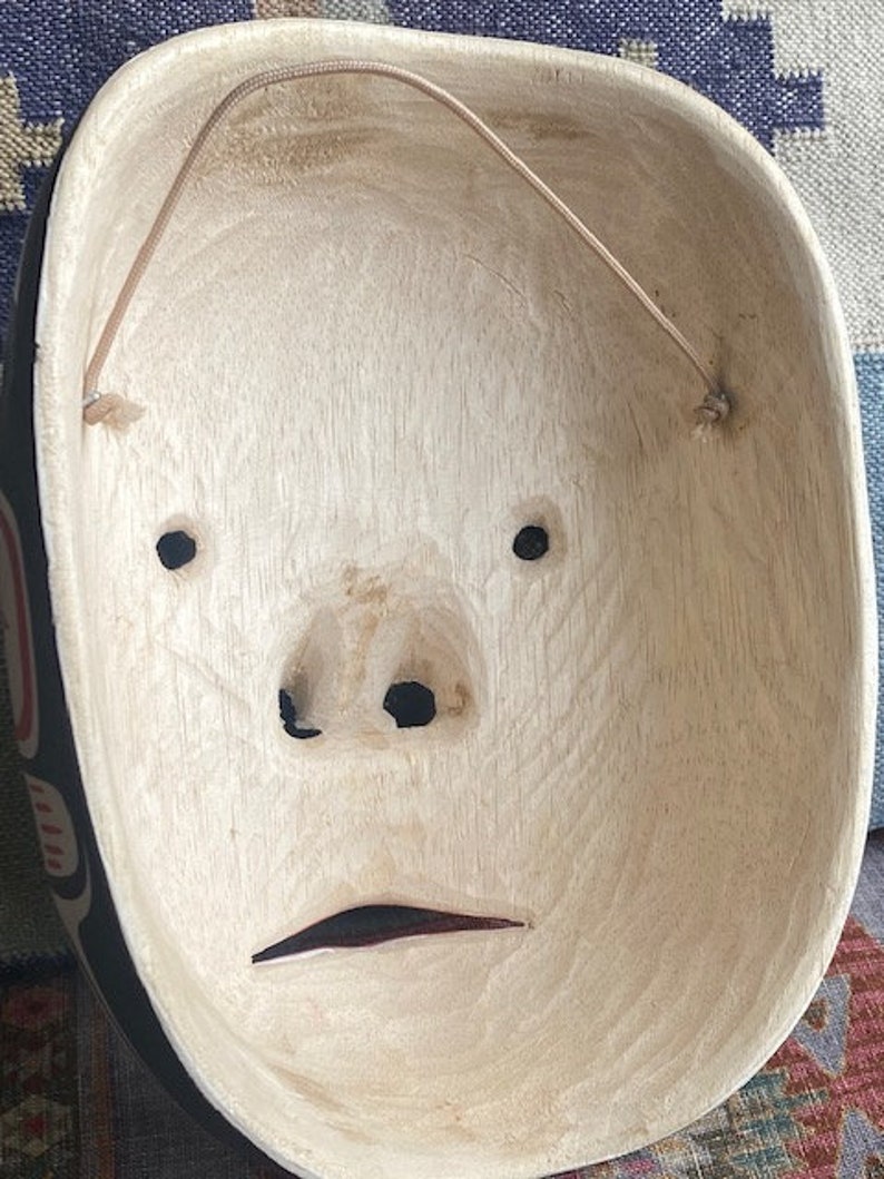 K&ouml;nnte beinhalten: Eine Holzmaske mit einem geschnitzten Gesicht und einer Schnur zum Aufh&auml;ngen. Die Maske ist mit schwarzen und roten Details bemalt.