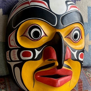 K&ouml;nnte beinhalten: Eine Holzmaske mit einem stilisierten menschlichen Gesicht, bemalt in Gelb, Schwarz, Rot und Wei&szlig;. Die Maske hat gro&szlig;e Augen, eine prominente Nase und einen breiten Mund.