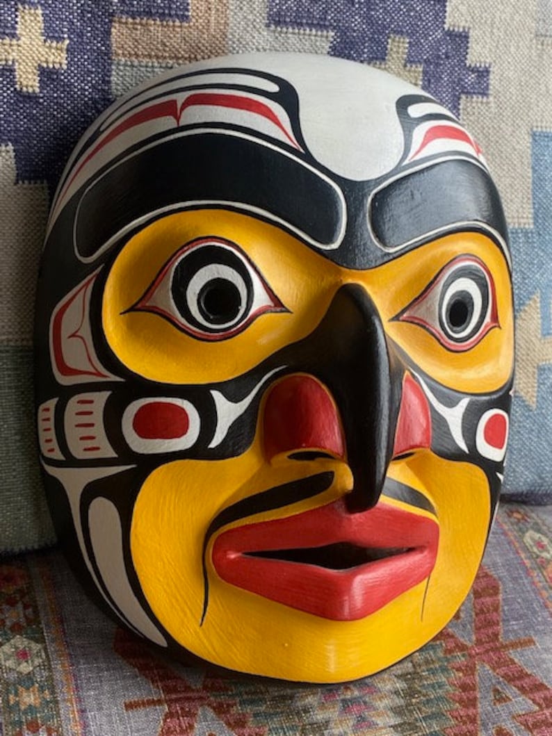 K&ouml;nnte beinhalten: Eine Holzmaske mit einem stilisierten menschlichen Gesicht, bemalt in Gelb, Schwarz, Rot und Wei&szlig;. Die Maske hat gro&szlig;e Augen, eine prominente Nase und einen breiten Mund.
