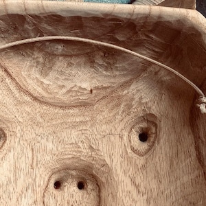 May include: A wooden bowl with a carved pig face design. The bowl has two holes for eyes and a snout with two nostrils. A white string is attached to the inside of the bowl.
