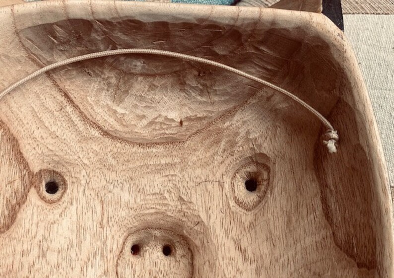 May include: A wooden bowl with a carved pig face design. The bowl has two holes for eyes and a snout with two nostrils. A white string is attached to the inside of the bowl.