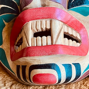 May include: A wooden mask with a carved mouth and teeth, painted in red, black, and green. The mask has a traditional design with geometric patterns.