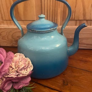 May include: A vintage blue enamel teapot with a curved handle and spout. The teapot has a gradient colour scheme, transitioning from light to dark blue. The lid is a lighter shade of blue. The teapot is placed on a wooden surface.