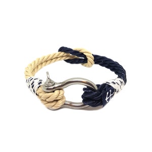 May include: A nautical rope bracelet with a stainless steel clasp. The bracelet is made of two colors of rope, one beige and one navy blue. The navy blue rope has a white anchor design.