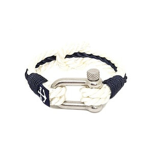 May include: White and navy blue rope bracelet with a silver anchor clasp. The bracelet is made of two strands of rope, one white and one navy blue, that are woven together. The clasp is a silver anchor with a small screw on the top.