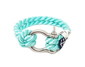 Puede incluir: Un brazalete de cuerda turquesa con un cierre de ancla de plata. El brazalete tiene un logotipo de ancla blanco y negro en el cierre.