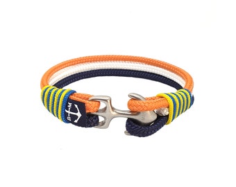 Op de afbeelding: Een nautische armband met oranje, witte en marineblauwe touwen. De armband heeft een roestvrijstalen sluiting en is geaccentueerd met blauw-gele gestreepte details. Een klein marineblauw label met een ankerlogo is zichtbaar.