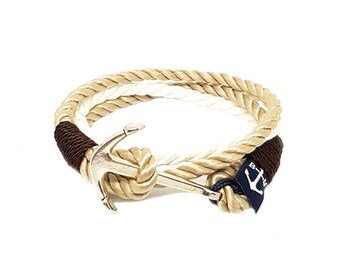 Op de afbeelding: Nautische armband met een zilveren ankerbedel en touwontwerp. De armband combineert beige, witte en bruine touwen met een donkerblauwe accent. Een klein label met een nautisch symbool is bevestigd.