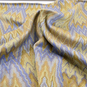 May include: Close-up of a woven fabric with a chevron pattern. The fabric features a mix of colors, including gold, light blue, and brown. The material is draped, showing the texture and weave of the fabric.