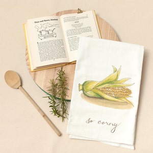 May include: A white kitchen towel with a watercolor illustration of an ear of corn. The text "so corny" is written below the corn.