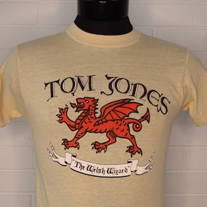 May include: Pale yellow t-shirt featuring the text "TOM JONES" above a red dragon graphic with the words "The Welsh Wizard" on a banner. The shirt is displayed on a mannequin against a white brick wall.