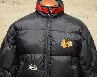 May include: Black puffer jacket with red accents and a Chicago Blackhawks logo on the chest. The jacket has a zip fastening and a high collar.