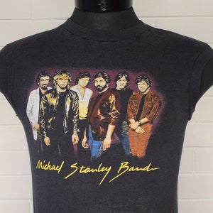 May include: Black sleeveless t-shirt with a colorful image of a band, the Michael Stanley Band, with the band's name in yellow lettering.