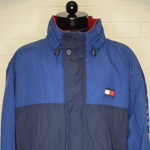 May include: A blue and navy blue jacket with a zipper closure and a Tommy Hilfiger logo on the chest. The jacket has a drawstring at the waist and a hood.