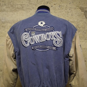 Peut inclure: Une veste de baseball bleue et beige avec le logo des Dallas Cowboys au dos. Le logo est brodé et indique "National Football League" et "Établi en 1960".