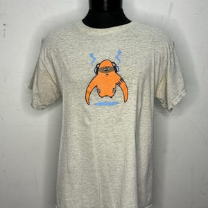 May include: A light gray t-shirt with a cartoon character wearing headphones and a black mask. The character is orange and has the word "SFR" written on its arm.