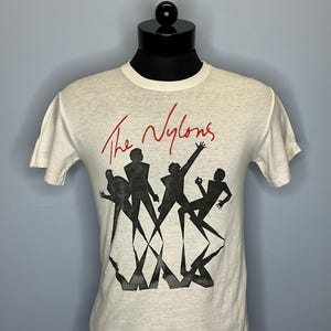 May include: White t-shirt with a black and white graphic of five figures in silhouette. The text "The Nylons" is written in red above the figures.