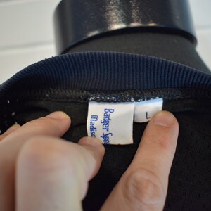 May include: Close-up of a black athletic shirt with a navy blue ribbed collar. The shirt has a tag that reads "Badger Sport Madison" and a size "L" label.