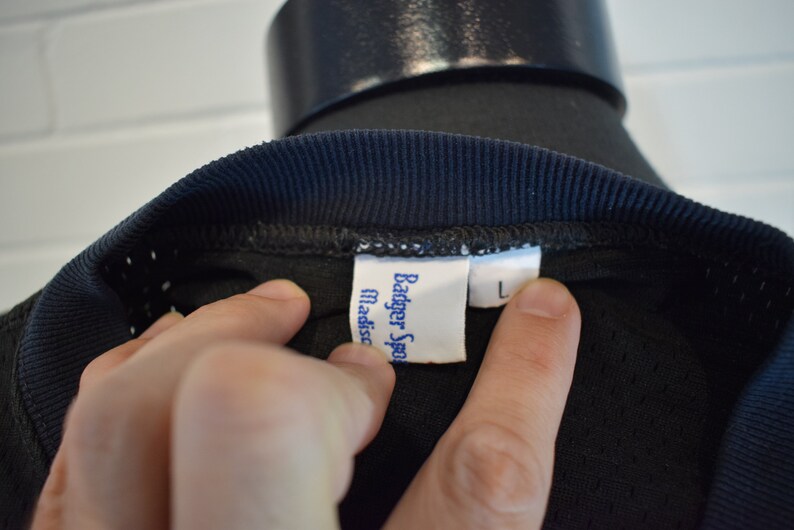 May include: Close-up of a black athletic shirt with a navy blue ribbed collar. The shirt has a tag that reads "Badger Sport Madison" and a size "L" label.