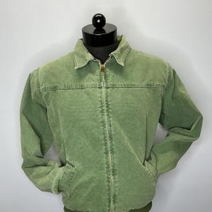 Puede incluir: Chaqueta verde con cremallera, dos bolsillos delanteros y cuello. La chaqueta tiene dobladillo y pu&ntilde;os acanalados. El logotipo de Carhartt es visible en la parte inferior derecha. La chaqueta se exhibe en una cabeza de maniqu&iacute; negra.