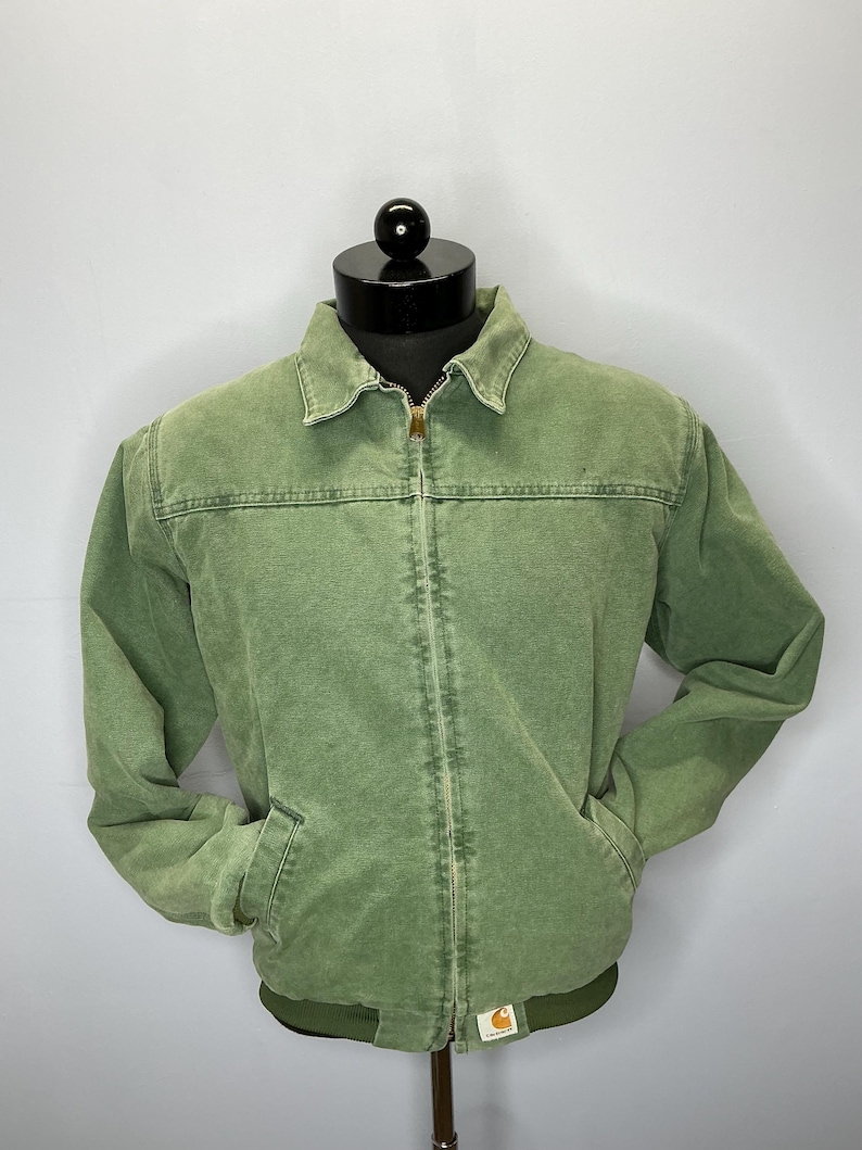 Puede incluir: Chaqueta verde con cremallera, dos bolsillos delanteros y cuello. La chaqueta tiene dobladillo y pu&ntilde;os acanalados. El logotipo de Carhartt es visible en la parte inferior derecha. La chaqueta se exhibe en una cabeza de maniqu&iacute; negra.