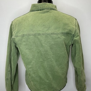 Puede incluir: Chaqueta verde con botones y mangas largas. La chaqueta tiene cuello y costura en la parte superior de la espalda. La tela tiene un aspecto descolorido. La chaqueta se exhibe en un maniqu&iacute;.