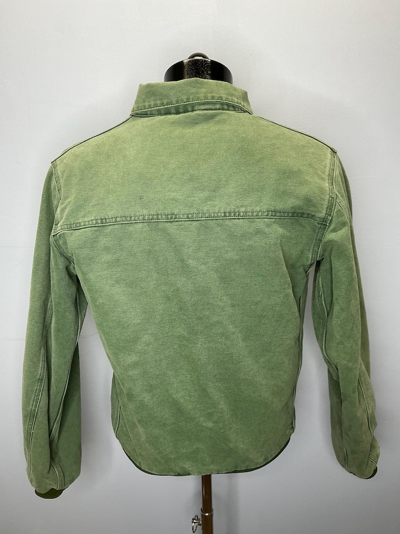 Puede incluir: Chaqueta verde con botones y mangas largas. La chaqueta tiene cuello y costura en la parte superior de la espalda. La tela tiene un aspecto descolorido. La chaqueta se exhibe en un maniqu&iacute;.