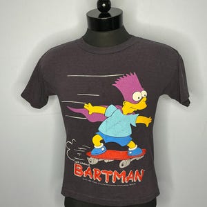 May include: Dark gray t-shirt featuring Bart Simpson as Bartman on a skateboard. The graphic includes Bart with purple hair, a blue shirt, and red skateboard. The word "BARTMAN" is in red and white.