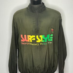 May include: Dark olive green windbreaker jacket with a half-zip and long sleeves. The front displays the text "SURF STYLE" in rainbow colours, with "Interplanetary Body Wear" below. A small logo is on the right chest.