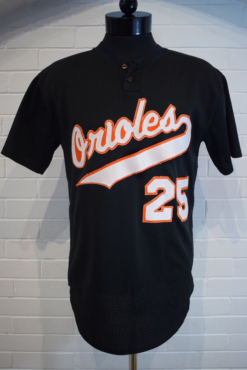 May include: Black baseball jersey featuring the word "Orioles" in orange and white script. The number "25" is displayed in orange and white on the front. The jersey has a button-up collar.