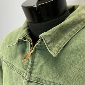 Puede incluir: Primer plano de una chaqueta verde con cremallera de lat&oacute;n. La chaqueta tiene cuello y detalles de costura visibles. La tela parece ser un material duradero, posiblemente lona. La chaqueta se exhibe en un maniqu&iacute; negro.