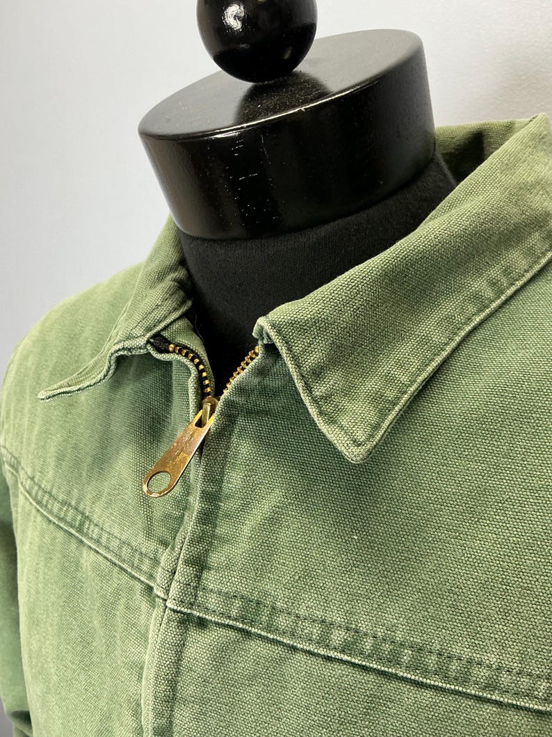 Puede incluir: Primer plano de una chaqueta verde con cremallera de lat&oacute;n. La chaqueta tiene cuello y detalles de costura visibles. La tela parece ser un material duradero, posiblemente lona. La chaqueta se exhibe en un maniqu&iacute; negro.