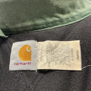 Puede incluir: Primer plano del cuello y la etiqueta de una chaqueta Carhartt. El cuello es verde oliva y el interior de la chaqueta es negro con un patr&oacute;n acolchado. La etiqueta muestra el logotipo de Carhartt y las instrucciones de lavado. Talla X-Large.