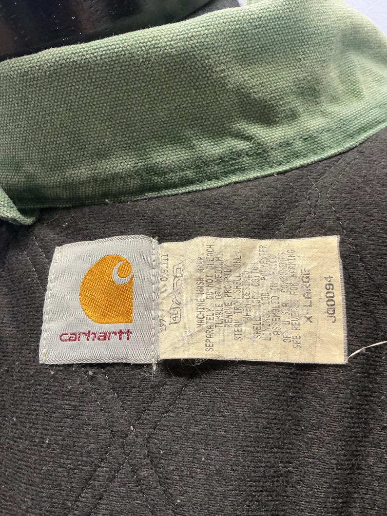 Puede incluir: Primer plano del cuello y la etiqueta de una chaqueta Carhartt. El cuello es verde oliva y el interior de la chaqueta es negro con un patr&oacute;n acolchado. La etiqueta muestra el logotipo de Carhartt y las instrucciones de lavado. Talla X-Large.